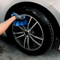 A hand wearing a black rubber glove holds a blue microfiber cloth, which is applying a tire shine product to a black tire with a complex silver alloy wheel. The tire has a high gloss finish and a few suds are visible at the bottom. Product used is Ultra Tire Shine.